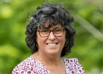 Janet, smiling, is a woman with dark, curly, short hair wearing glasses