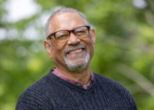 Nolan, smiling, is an African American man with facial hair and glasses wearing a blue sweater