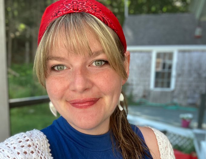 Savannah has blonde hair pulled back in a bandana, gold earrings and a blue shirt; she is smiling