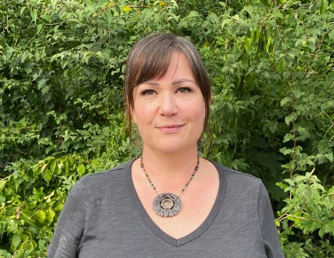 Kristin has short brown hair and bangs and is wearing a necklace and gray tshirt