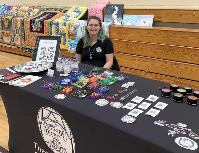 Ambrosia Ross sits at her table covered with branded merchandise to invite fair-goers to learn about Theater of Dice