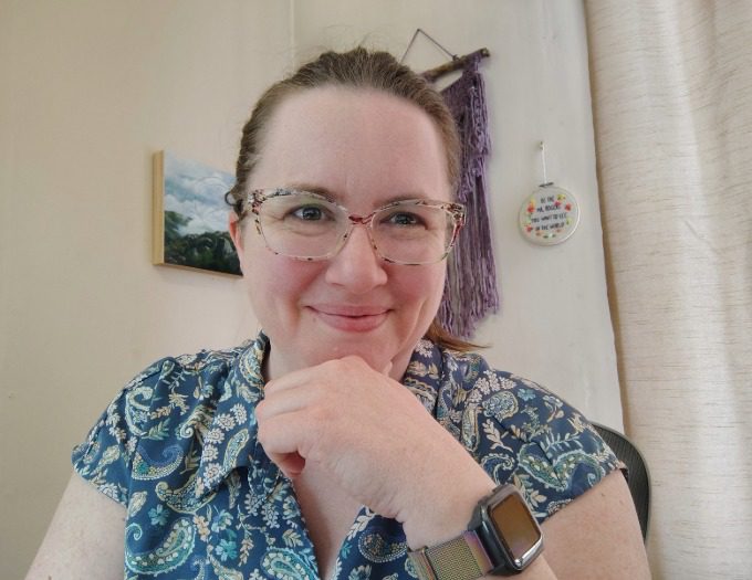 Victoria Flynn wears a blue foral pattern shirt, her brown hair back and has glasses and is smiling at the camera