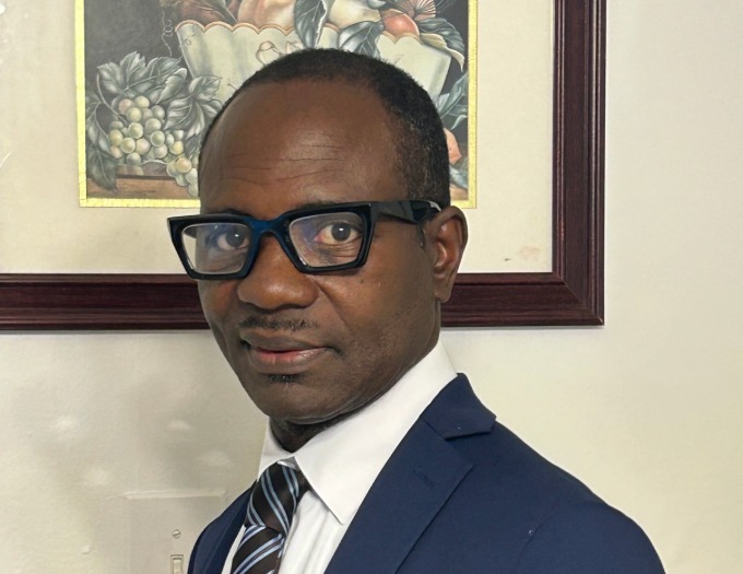 Victorien wears black-rimmed glasses and a suit and tie. He is standing in front of a floral painting.
